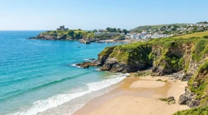 A sunny sandy beach cove in South Cornwall with turquoise waves, green sea cliffs, a coastal village, and a historic castle on a distant headland